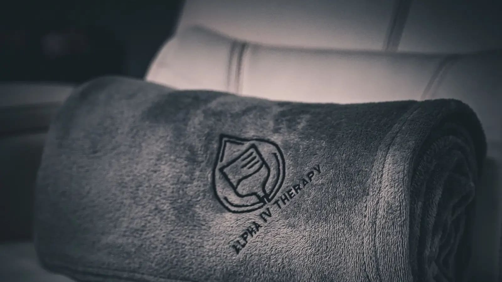 Rolled gray blanket embroidered with the Alpha IV Therapy logo on an infusion chair